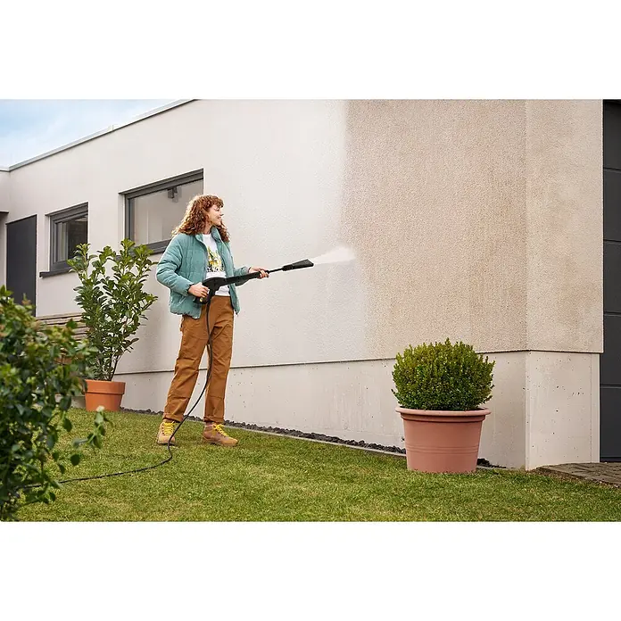Kärcher Strahlrohr eco!Booster 120 - Passend für: Kärcher Hochdruckreiniger K 2 und K 3, 452 x 104 x 42 mm - Eine Frau reinigt eine schmutzige Hauswand mit einem schwarz-gelben Hochdruckreiniger, der einen sauberen Streifen hinterlässt.