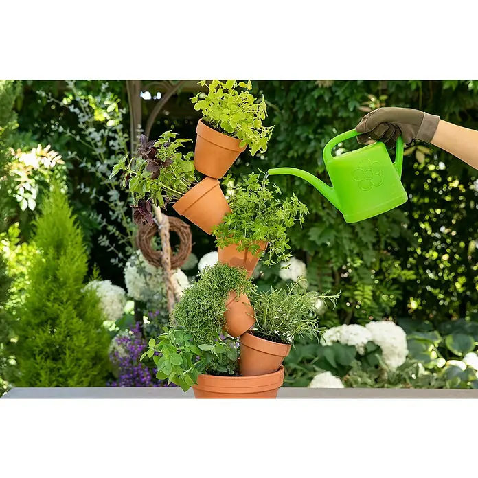 Piardino Kräuterpflanzen-Mix Mediterran - 4 Stk., verschiedene Sorten, Topfgröße: 12 cm - Kreativer Kräutergarten mit gestapelten Tontöpfen und einer grünen Gießkanne, die die Pflanzen bewässert, vor einem üppigen Gartenhintergrund.
