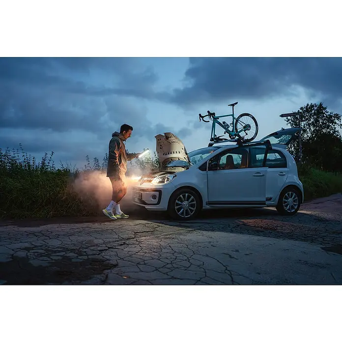 Ledlenser LED Baustrahler AF2R WORK - 1.000 lm, 6.000 K - 7.000 K, 4,6 x 3,8 x 24,8 cm, IP54 - Mann beleuchtet mit einer tragbaren Lampe den Motor eines rauchenden Autos bei Nacht. Ein Fahrrad ist auf dem Dachgepäckträger.