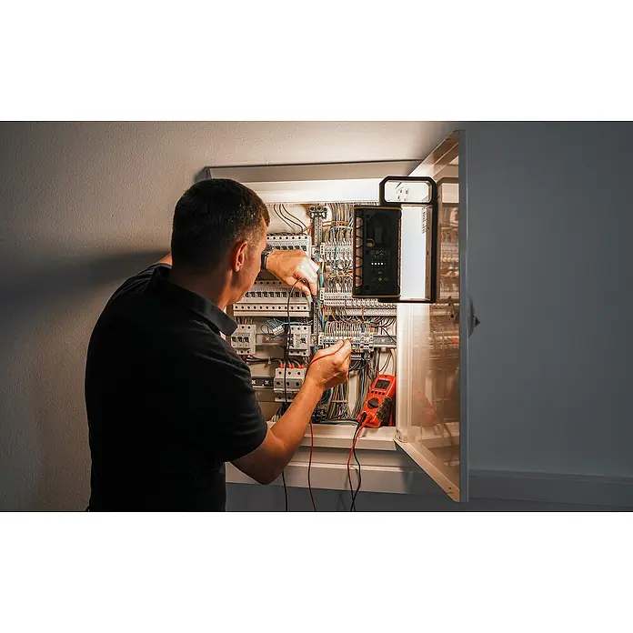 Ledlenser LED Baustrahler AF4R WORK - 2.000 lm, 3.000 K - 6.500 K, 4,4 x 26,6 x 11,9 cm, IP67 - Elektriker bei der Fehlersuche an einer Schalttafel mit rotem Multimeter und Arbeitsleuchte.