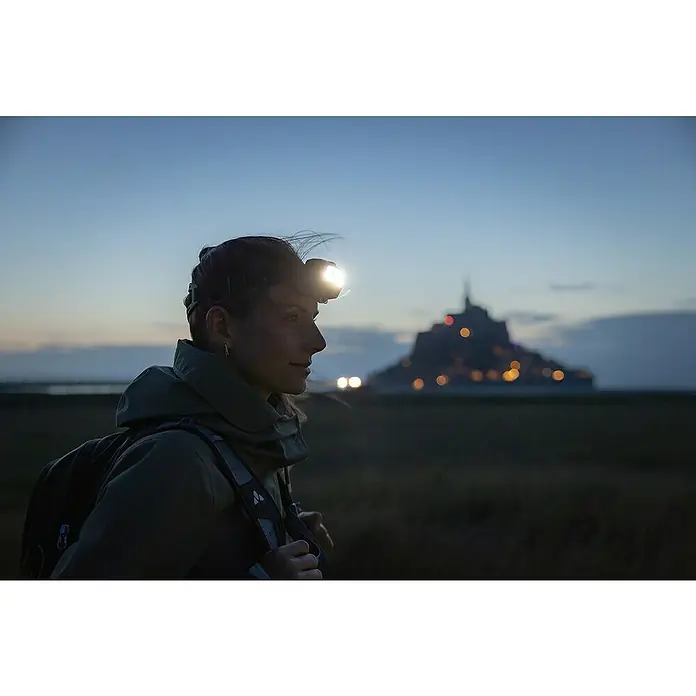Ledlenser Stirnlampe HF6R Core - 800 lm, IP68 - Frau mit leuchtender Stirnlampe und Rucksack wandert bei Dämmerung draußen, Mont Saint-Michel im Hintergrund.