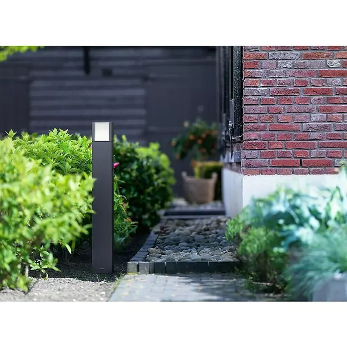 Philips Sobremuro exterior Arbour - Color del marco: Negro, Blanco frío, 10 x 10 x 77 cm, Con bombillas - Lámpara moderna de exterior de metal con un cabezal rectangular luminoso, colocada en un jardín entre plantas y un camino de piedra.