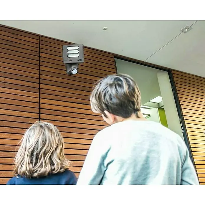Lutec Proyector LED Esa con sensor y cámara - 22 W, L x An x Al: 17 x 20 x 18 cm, Gris, Blanco frío - Una luz solar con sensor de movimiento está montada en una pared de madera. Dos personas son visibles en el fondo.