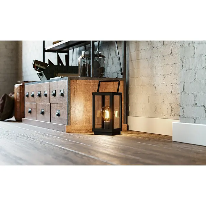 Eglo Deko-Tischleuchte BRADFORD 160 W, L x B x H: 16,5 x 15 x 37 cm, Schwarz, E27 - Nahaufnahme einer eleganten schwarzen Laterne mit einer Edison-Glühbirne, die auf einem Holzboden vor einem rustikalen Schrank und einer Ziegelwand steht.