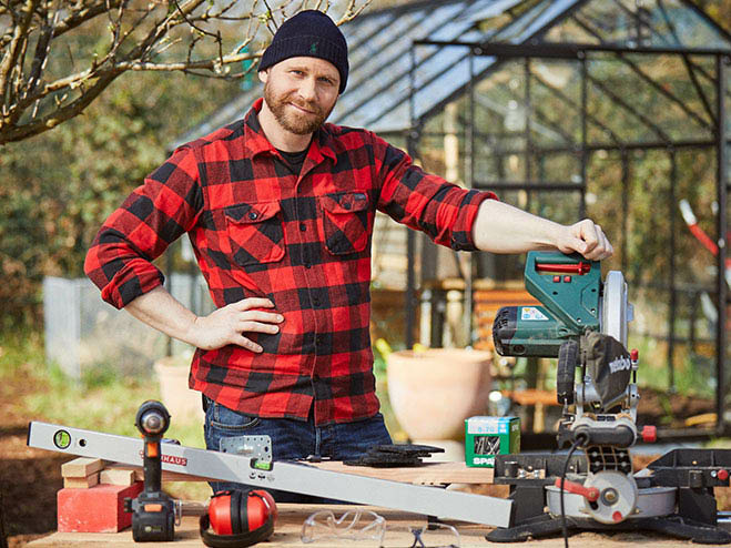 Mann steht im Garten vor einem Tisch mit verschiedenen Materialien wie einer Säge für den DIY Gartenweg