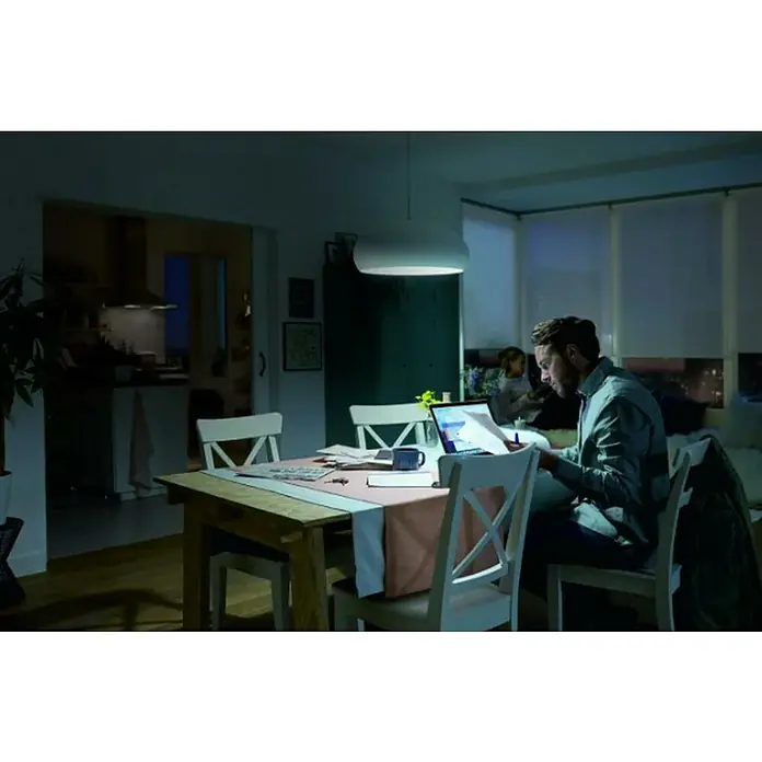 Philips Bombilla LED Vela y lustre CDL - E14, No regulable, Blanco diurno, 470 lm, 40 W - Un hombre está sentado a la mesa trabajando en su computadora portátil. La lámpara colgante blanca ilumina el espacio para trabajar y estudiar.