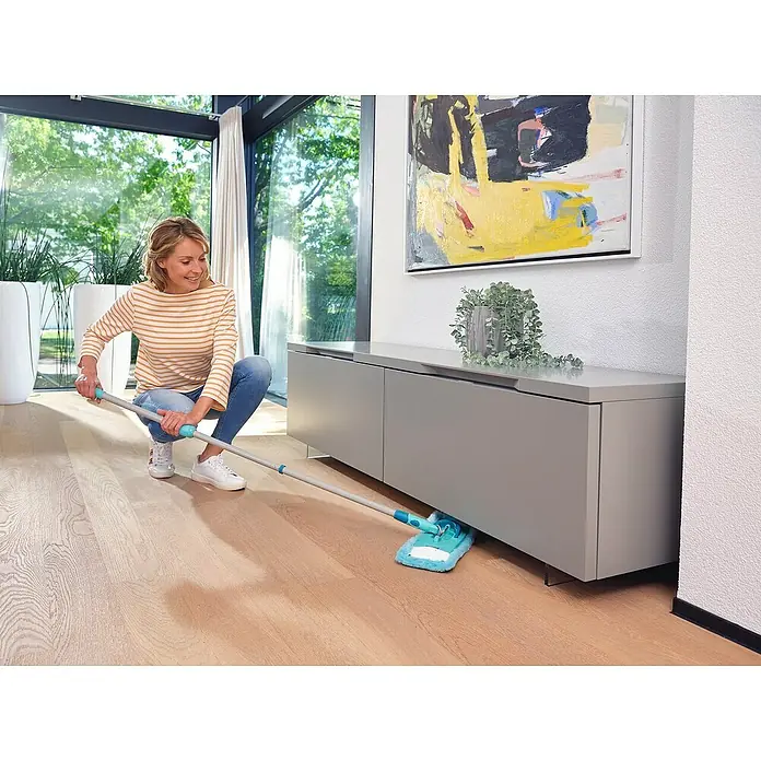 Leifheit Bodenwisch-Set Clean Twist M Ergo - 4 -tlg., 6 l - Eine Frau putzt mit einem blauen Mopp unter einem grauen Schrank. Der Mopp hat einen langen Griff und die Frau trägt ein gestreiftes Oberteil.