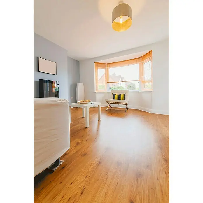 Suelo vinílico autoadhesivo Roble natural - 91,4 cm x 15,2 cm x 2 mm, Marrón - Sala de estar con suelo laminado, sofá, mesa de centro, sillón y ventana con persianas. Un ambiente luminoso y acogedor perfecto para relajarse.