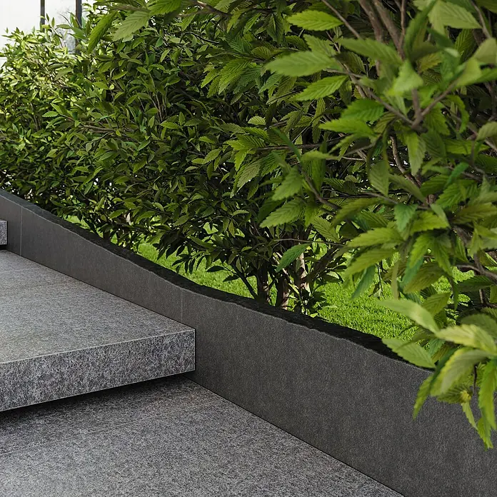 Rasenkante - Anthrazit, Basalt - Eine nahaufnahme der Steintreppe, die zu einem Garten mit grünen Büschen führt. Die Steinstufen haben eine raue, dunkle Oberfläche.