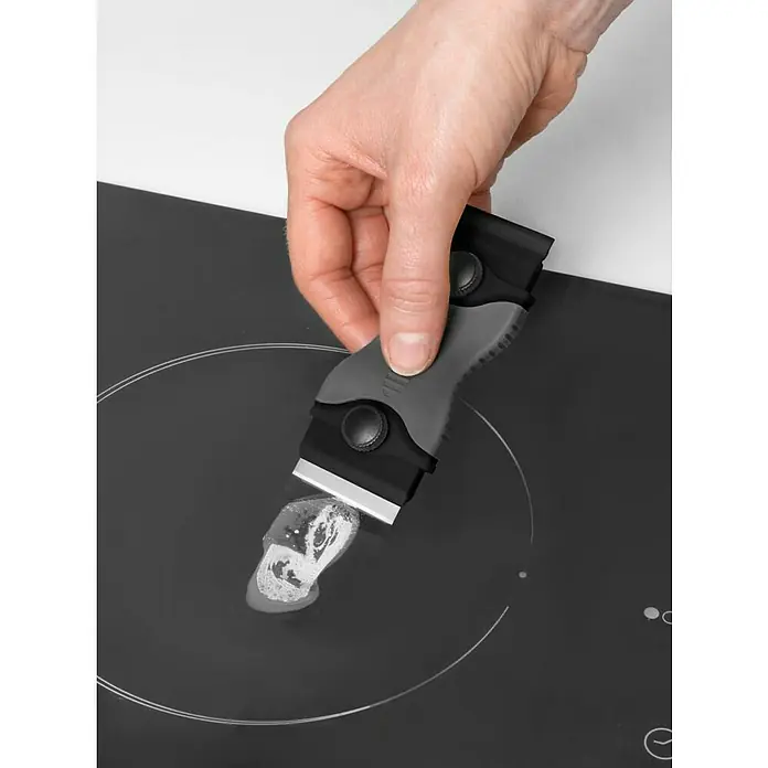 Glasschaber - Passend für: Für alle glatten Oberflächen - Hand reinigt mit einem dunkelgrauen und schwarzen Schaber mit Metallklinge weiße Rückstände von einem schwarzen Induktionskochfeld.
