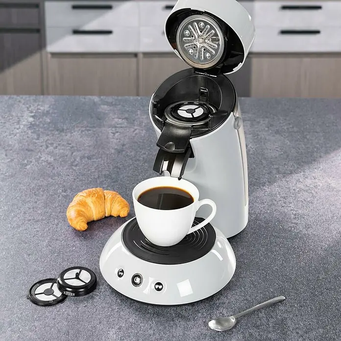 Xavax Kaffeepad - 2 Stk. - Weiße Kaffeekapselmaschine brüht Kaffee in Tasse auf Küchentheke, dazu Croissant. Deckel offen, Innenleben sichtbar.