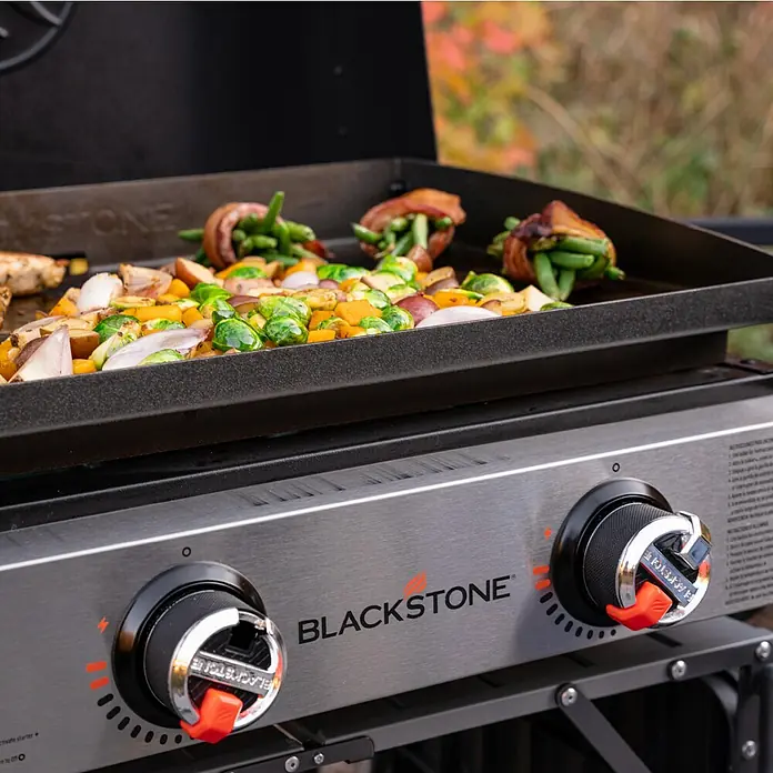 Blackstone Plancha Grill 22" - Superficie de la parrilla (An x Pr): 57 x 41,5 cm, Negro, Número de quemadores: 2 ud. - Plancha Blackstone mostrando verduras de otoño y atados envueltos en tocino, ideal para cocinar al aire libre y comidas deliciosas.
