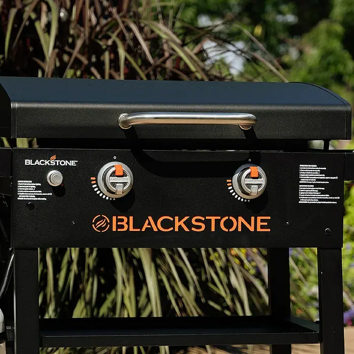 Blackstone Plancha-Grill 28" mit Haube - Grillfläche (B x T): 73 x 46,3 cm, 10 kW, Schwarz - Nahaufnahme eines schwarzen Blackstone Grills mit zwei Reglern und einem Edelstahlgriff vor Pflanzen.