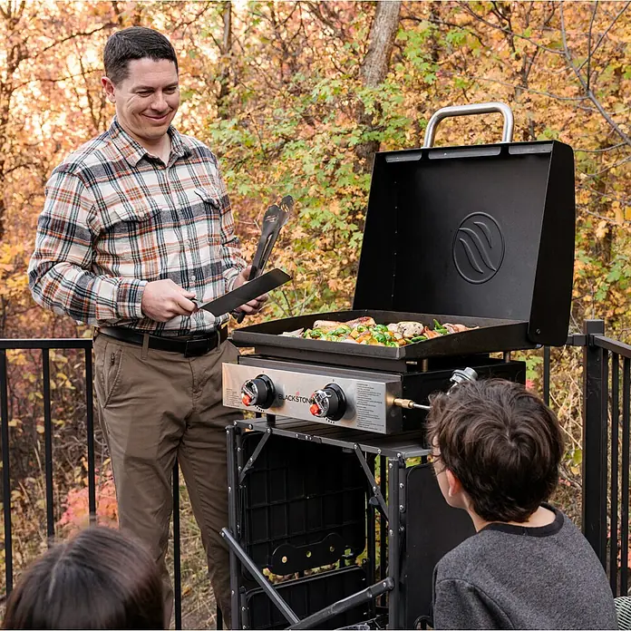 Blackstone Plancha-Grill 22" mit Haube - Grillfläche (B x T): 57 x 41,5 cm, 7 kW, Schwarz - Ein Mann grillt auf einem schwarzen Blackstone-Grill im Freien, während Kinder zuschauen. Perfekt für Outdoor-Kochabenteuer!