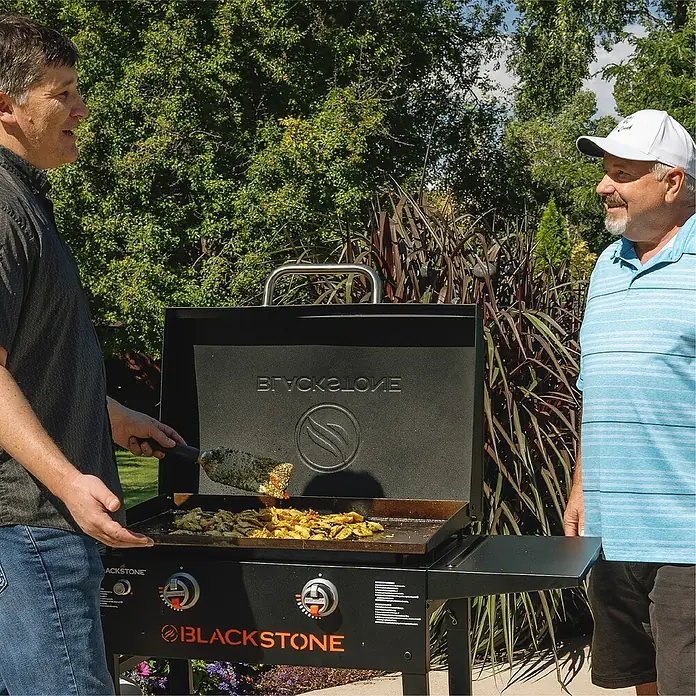 Blackstone Plancha-Grill 28" mit Haube - Grillfläche (B x T): 73 x 46,3 cm, 10 kW, Schwarz - Zwei Männer stehen an einem Blackstone Griddle, auf dem Essen zubereitet wird. Der Grill steht im Freien, umgeben von Pflanzen.
