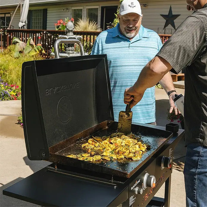 Blackstone Plancha-Grill 28" mit Haube - Grillfläche (B x T): 73 x 46,3 cm, 10 kW, Schwarz - Zwei Männer bereiten auf einem schwarzen Blackstone-Grill im Freien eine Mahlzeit zu, wobei die offene Grillplatte mit gebratenem Gemüse bedeckt ist.