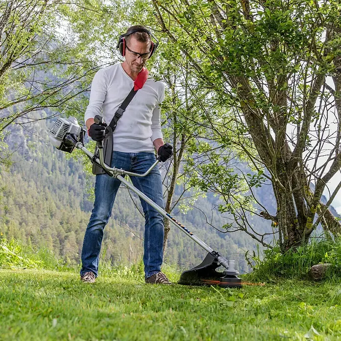 AL-KO Benzin-Motorsense BC 223 B - 700 W, Schnittbreite Faden: 41 cm, Schnittbreite Messer: 25 cm - Der Trimmer im Einsatz: Ein Mann mäht Gras mit einem Rasentrimmer, geschützt mit Brille und Kopfhörern, für präzise Gartenarbeit.