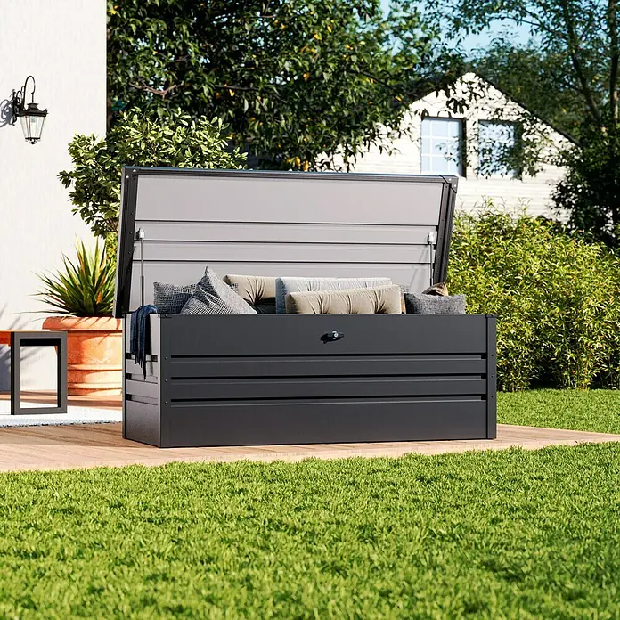 Hoggar Garten-Aufbewahrungsbox Sveg - L x B x H: 165 x 70 x 62 cm, Anthrazit - Eine geöffnete, graue Gartenbox mit Kissen und Decken, die auf einer Holzterrasse im Grünen steht. Sie bietet viel Stauraum.