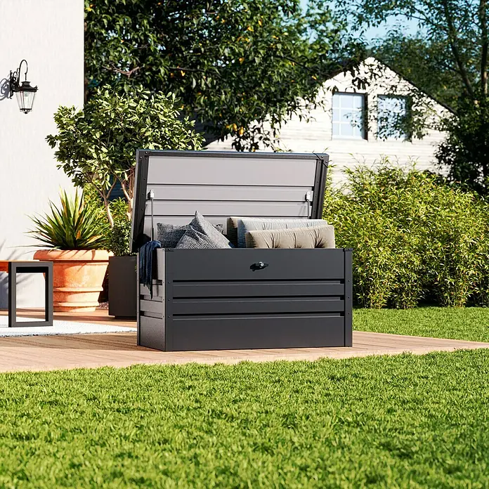 Hoggar Garten-Aufbewahrungsbox Sveg - L x B x H: 120 x 61 x 62 cm, Anthrazit - Eine graue Aufbewahrungsbox mit Kissen und Decke steht auf einer Holzterrasse im Garten. Die Box bietet Stauraum für Gartenutensilien.
