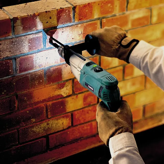 Makita Schlagbohrmaschine HP2071FJ - 1.100 W, Schnellspannbohrfutter - Ein Handwerker bohrt mit einer Makita Schlagbohrmaschine ein Loch in eine Ziegelwand, wobei der Bohrer und die Maschine im Fokus stehen.