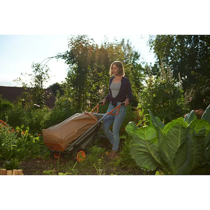 Worx Carretilla de obra Aerocart multifunción 8 en 1 - 75 l, Acero, Capacidad de carga: 136 kg - Mujer empujando un carro de jardín gris con asas y ruedas naranjas, cubierto por una lona marrón, en un jardín.