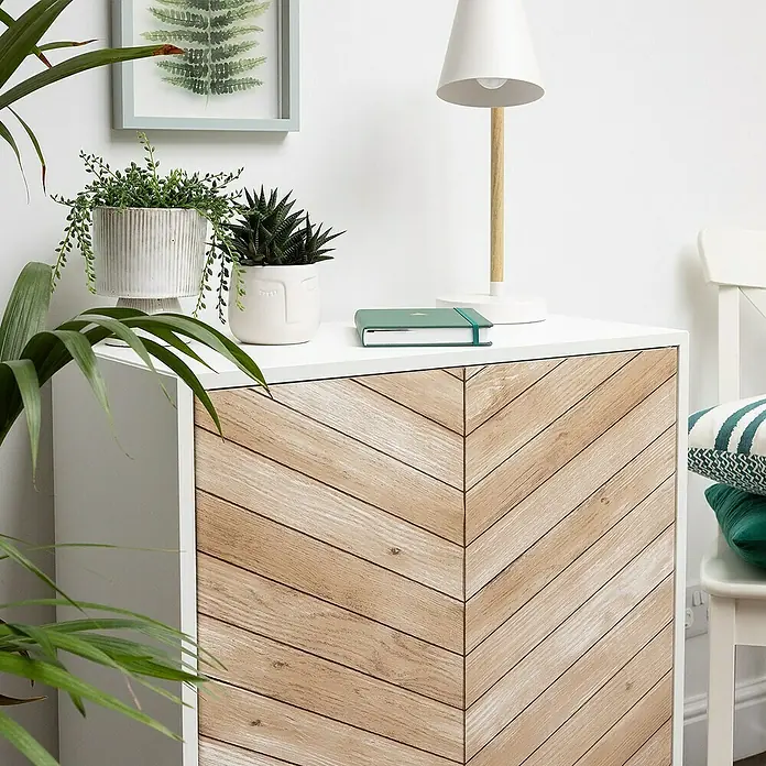 D-c-fix Rollo adhesivo vinílico Espiga Madera - 200 x 45 cm, Marrón, Autoadhesivo - Una elegante cómoda con un patrón de espiga en los cajones, decorada con plantas y una lámpara.
