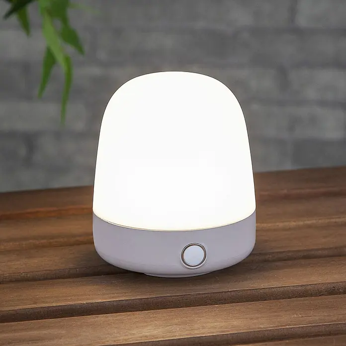 Starlux Akku-Tischleuchte Mini - L x B x H: 10 x 10 x 12 cm, Beige, RGBW, 0,3 W, IP44 - Nahaufnahme einer strahlenden, kleinen Tischlampe mit weißem Lampenschirm und grauem Fuß auf einem Holztisch.