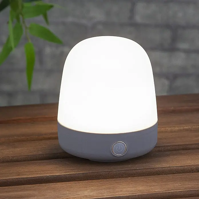 Starlux Akku-Tischleuchte Mini - L x B x H: 10 x 10 x 12 cm, Light Grey, RGBW, 0,3 W, IP44 - Eine graue LED-Tischleuchte mit einem runden Power-Button steht auf einer braunen Holzoberfläche und spendet warmes, diffuses Licht.