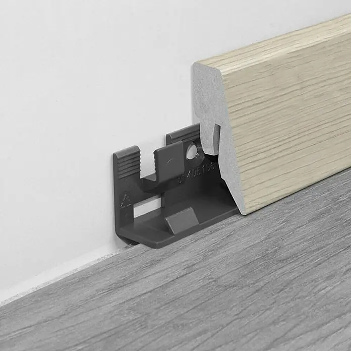 LOGOCLIC Sockelleiste K40 C Steirische Eiche - 260 cm x 19 mm x 40 mm, MDF - Graue Sockelleistenbefestigung aus Kunststoff mit einer hellen Holzleiste an einer weißen Wand und einem grauen Boden.