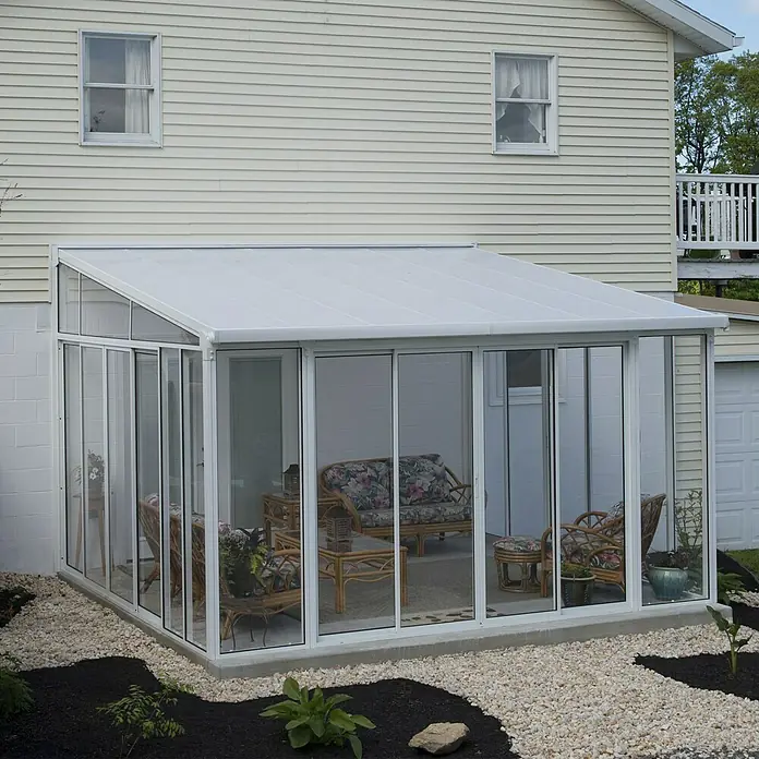 Palram – Canopia Wintergarten San Remo - B x T x H: 4,25 x 4 x 3,1 m, Verkehrsweiß - Außenansicht eines Wintergartens mit weißen Rahmen und viel Glas, der an ein Haus mit hellbeiger Fassade angebaut ist.