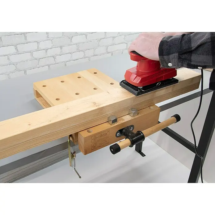 Rikta Hobelbank - L x B: 47 x 29 cm, Arbeitshöhe: 7 cm, Holz - Nahaufnahme eines Werkstatttisches mit Schraubstock, auf dem ein Mann mit einem roten Schleifgerät ein Holzbrett bearbeitet.