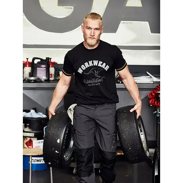 Terrax Workwear T-Shirt - XXL, Gelb/Schwarz - Ein muskulöser Mann in Roadsign Workwear trägt Reifen in einer Werkstatt, präsentiert robuste und stilvolle Arbeitskleidung.