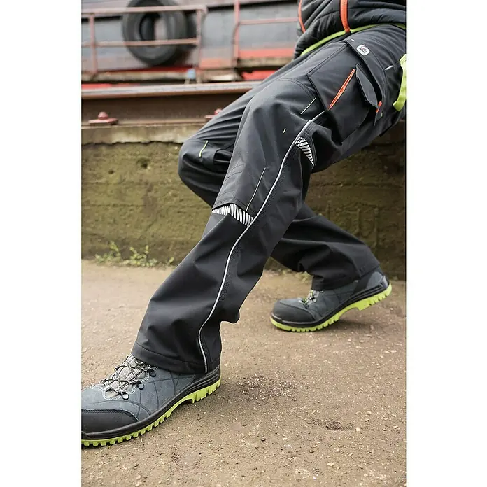 Terrax Workwear Herren-Arbeitshose - 52, Schwarz/Limette - Nahaufnahme der schwarzen Arbeitshose mit Taschen und Reflexionsdetails, kombiniert mit robusten Arbeitsschuhen mit hellgrüner Sohle.