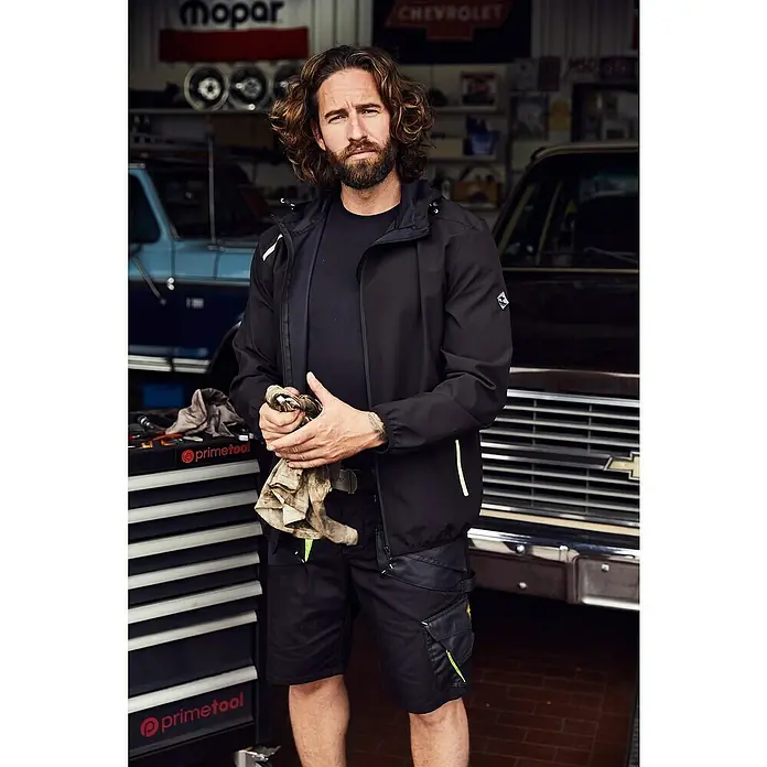 Terrax Workwear Arbeitsjacke - M, Schwarz/Limette - Ein Mann mit Bart präsentiert eine schwarze Jacke und Shorts der Marke primetool. Perfekt für die Werkstatt!