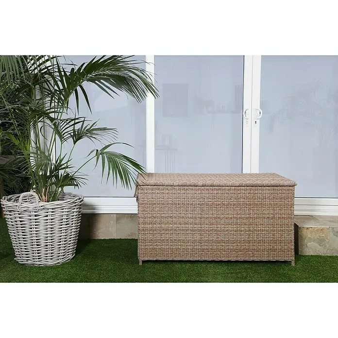 Baúl para jardín Gotland - 150 x 65 x 68 cm, Plástico, Arena - Una espaciosa caja de almacenamiento de ratán, ideal para guardar utensilios de jardín y cojines, se coloca junto a una planta en maceta en el césped.