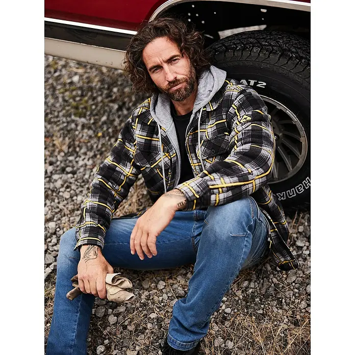 Terrax Workwear Thermojacke - M, Anthrazit/Gelb - Lässig sitzender Mann mit karierter Flanelljacke mit Kapuze, Jeans und Handschuhen vor einem roten Truck.