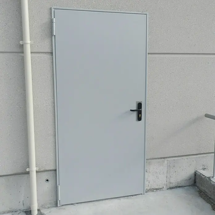 Novoferm Puerta cortafuegos EI2 60 C5 - 88,5 x 203,5 cm, Derecha, Blanco - Puerta exterior gris con manija negra sobre una pared de hormigón. La puerta ofrece un acceso seguro y duradero.