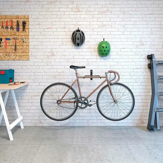 Rei Fahrrad-Wandhalter - L x B x H: 9 x 32 x 45 cm, Stahl, Wandmontage - Schwarze Fahrrad-Wandhalterung hält kupferfarbenes Rennrad an weißer Ziegelwand. Helme und Werkzeuge sind sichtbar.