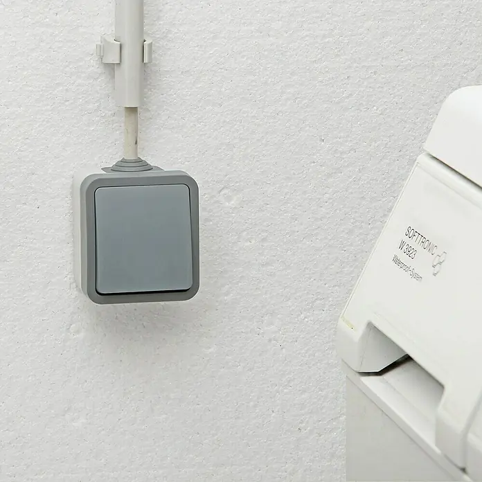 Voltomat Blueline Feuchtraumtaster - Grau, Aufputzmontage, IP54 - Nahaufnahme eines wasserdichten SOFTTRONIC W 3923 Schalters mit grauem Knopf und weißem Gehäuse an einer weißen Wand.