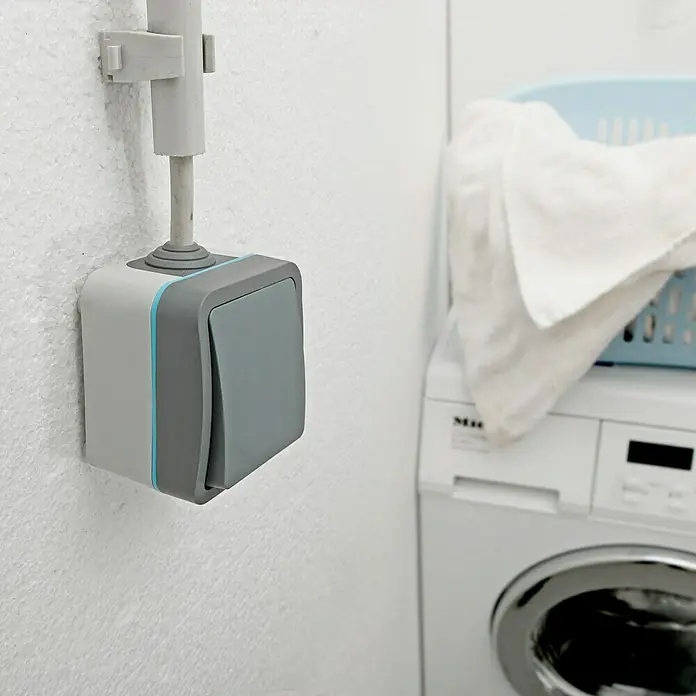 Voltomat Blueline Feuchtraumtaster - Grau, Aufputzmontage, IP54 - Nahaufnahme eines grauen Lichtschalters an einer Wand, der sich neben einer Waschmaschine in einem hellen Raum befindet.