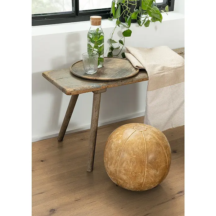 Egger Laminat NatureSense - Wildeiche Natur, Landhausdiele, 1.292 x 193 x 7 mm - Vintage-Szene mit rustikalem Holzhocker, Dekanter, Glas und einem Lederball, die eine gemütliche und nostalgische Atmosphäre schaffen.
