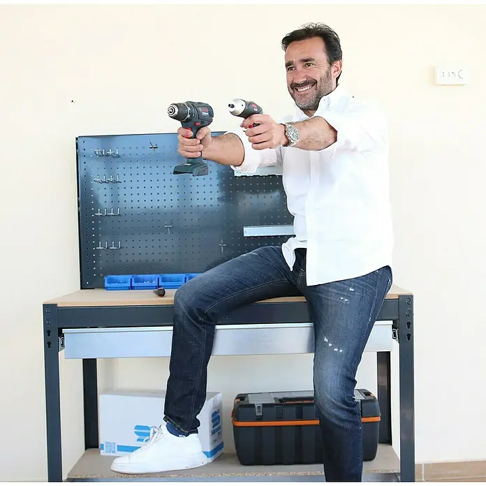 Simonrack Simonwork Banco de trabajo BT2 Box - Azul, Ancho: 121 cm - Banco de trabajo gris oscuro con panel perforado, un hombre se sienta sonriendo, sosteniendo dos taladros.