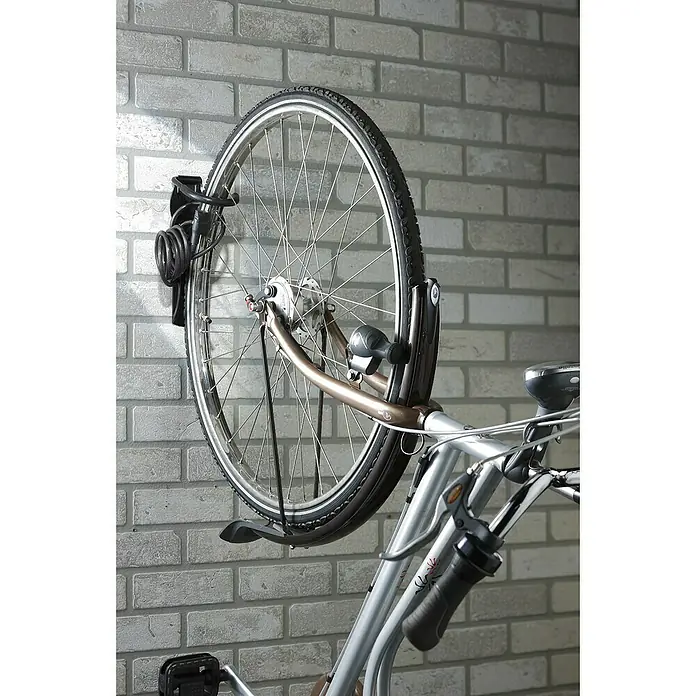 Mottez Gancho de pared para bicicletas B865V - Acero, Negro - Bicicleta plateada con horquillas de bronce montada verticalmente en una pared de ladrillo claro con un soporte negro. Un candado de cable está en la rueda.