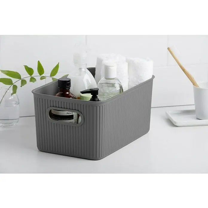 Tatay Baobab Cesta para la ropa - 18,1 x 27,2 x 13 cm, Plástico, Gris - Una caja de almacenamiento de plástico acanalada gris llena de artículos de baño como toallas, lociones y una planta.