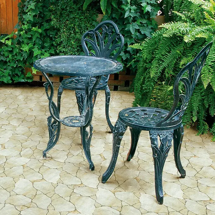 Feinsteinzeugfliese Cala Jet - 45 x 45 cm, Beige, Matt - Elegantes Gartenmöbelset mit einem runden Tisch und zwei Stühlen aus dunklem Metall, umgeben von üppigem Grün.