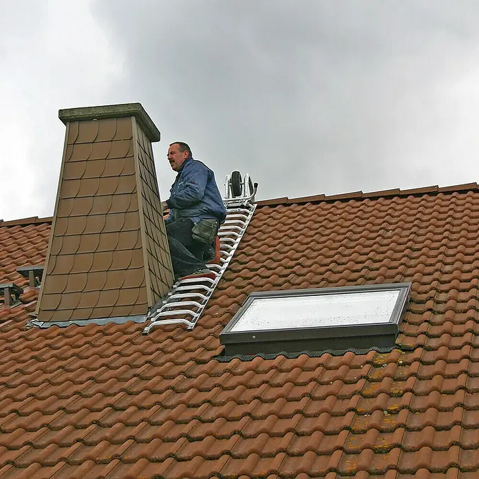 Krause First-Überwurfbügel - L x B x H: 1,1 x 0,5 x 0,6 m, Geeignet für: Dachleitern - Ein Mann auf einer Leiter auf einem Ziegeldach, der einen Schornstein inspiziert oder repariert. Eine Dachluke ist ebenfalls sichtbar.