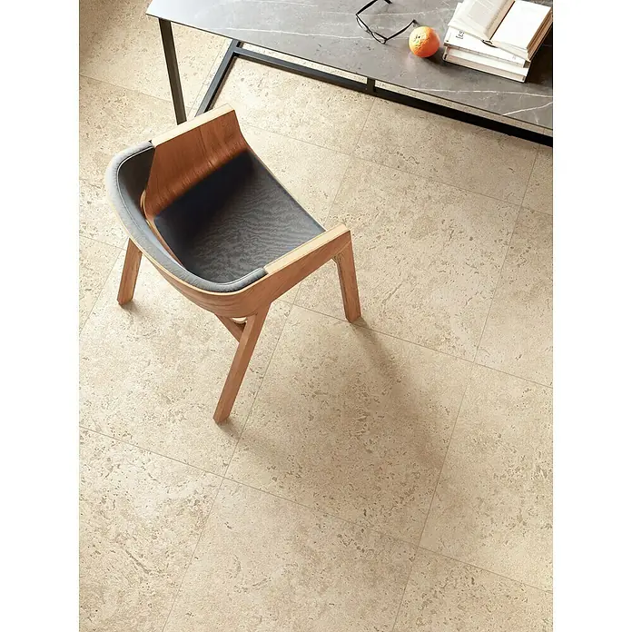 Corso Italia Terrassenfliese Pompei Aureo Crosscut - 60 x 60 cm, Beige, Matt - Blick von oben auf einen Stuhl aus Holz und grauem Stoff, der auf einem Fliesenboden steht, neben einem Tisch mit Büchern und Obst.