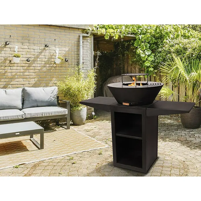 Purline Barbacoa de carbón EFP57B - Diámetro de la superficie de la parrilla: 60 cm, Negro - Esta imagen muestra una mesa de hoguera negra moderna con llamas, ubicada en un patio pavimentado rodeado de vegetación exuberante.