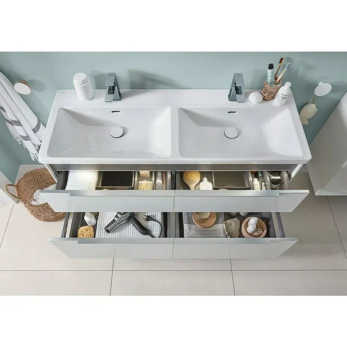 Villeroy & Boch Subway 3.0 Waschbeckenunterschrank - Brilliant White, Anzahl Schubladen: 4 Stk., Farbe Griff: Aluminium glänzend, B x T x H: 127,2 x 47,8 x 57,6 cm, Mit Beleuchtung, Geeignet für Doppelwaschtisch - Weißer Doppelwaschtisch-Badezimmerschrank mit geöffneten Schubladen, die organisierte Toilettenartikel, einen Föhn und hellgraue Aufbewahrungseinsätze zeigen.
