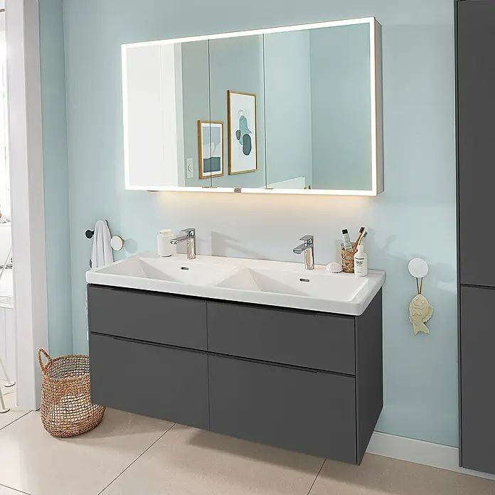 Villeroy & Boch Subway 3.0 Waschbeckenunterschrank - Graphite, Anzahl Schubladen: 4 Stk., Farbe Griff: Volcano Black, B x T x H: 127,2 x 47,8 x 57,6 cm, Mit Beleuchtung, Geeignet für Doppelwaschtisch - Ein moderner dunkelgrauer Doppelwaschtisch mit zwei weißen Waschbecken und beleuchtetem Spiegelschrank im hellblauen Bad.